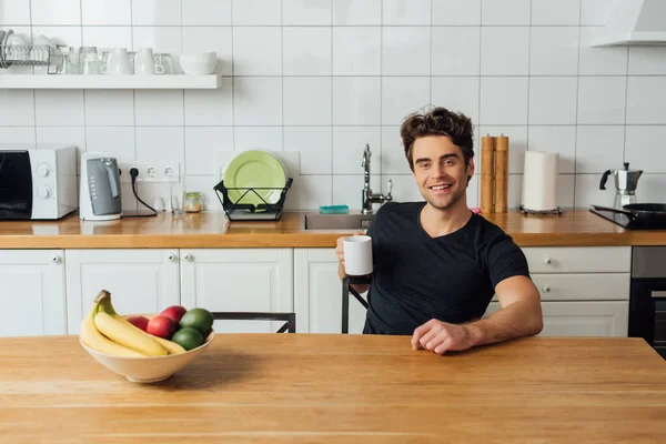 Selektiver Fokus eines gutaussehenden Mannes mit einer Tasse Kaffee, der in der Küche in die Kamera lächelt — Stockfoto