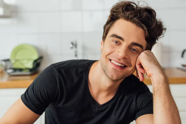 Bonito homem com a mão perto da bochecha sorrindo para a câmera em casa — Fotografia de Stock