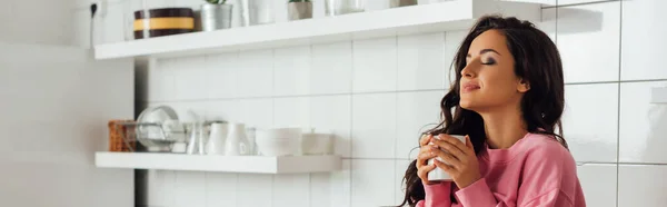 Tiro panorâmico de menina bonita com olhos fechados segurando xícara de café na cozinha — Fotografia de Stock