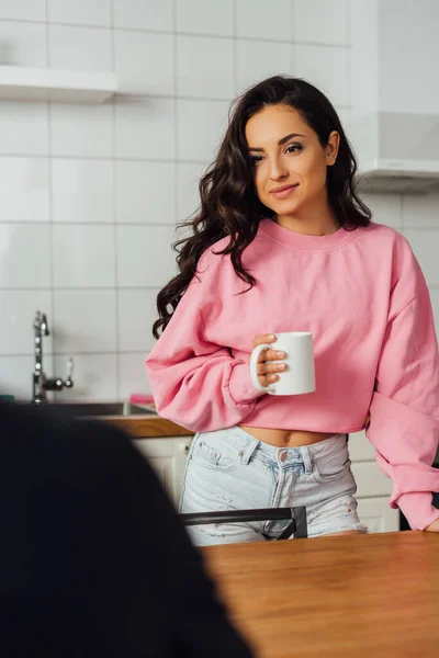 Selective focus of beautiful brunette girl holding cup of coffee near table in kitchen — Stock Photo