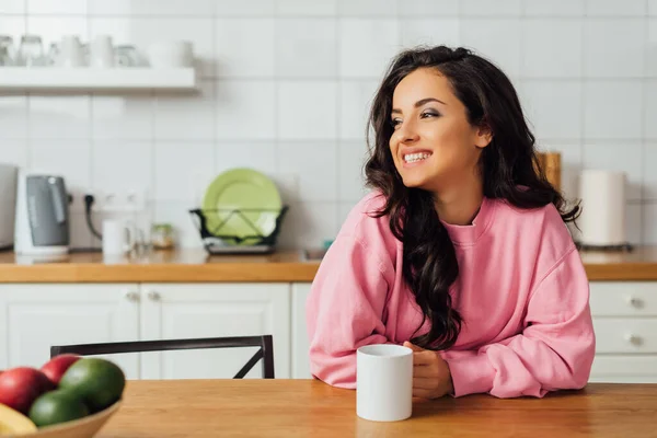 Foco seletivo da bela mulher morena sorrindo e segurando xícara de café à mesa na cozinha — Fotografia de Stock