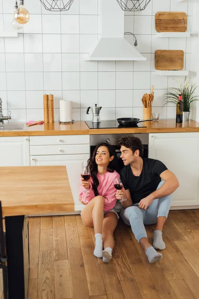 Selective focus of handsome man with glass of wine looking at smiling girlfriend on floor in kitchen — Stock Photo
