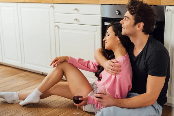 Sonriente hombre abrazando atractiva chica con vaso de vino mientras está sentado en el suelo en la cocina - foto de stock
