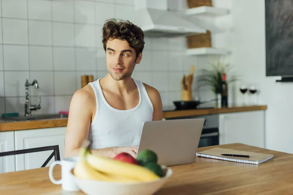 Enfoque selectivo de freelancer guapo usando portátil cerca de portátil y taza de café en la mesa de la cocina - foto de stock