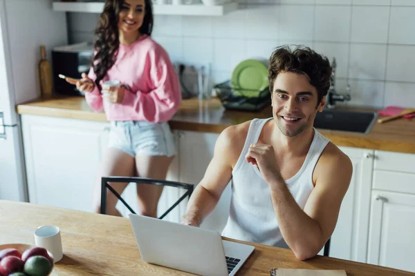 Enfoque selectivo del hombre sonriente utilizando el ordenador portátil cerca de la novia con taza y teléfono inteligente en la cocina - foto de stock