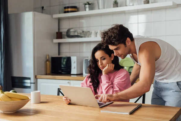 Selective focus of man pointing with finger at laptop near girlfriend and notebook on kitchen worktop — Stock Photo