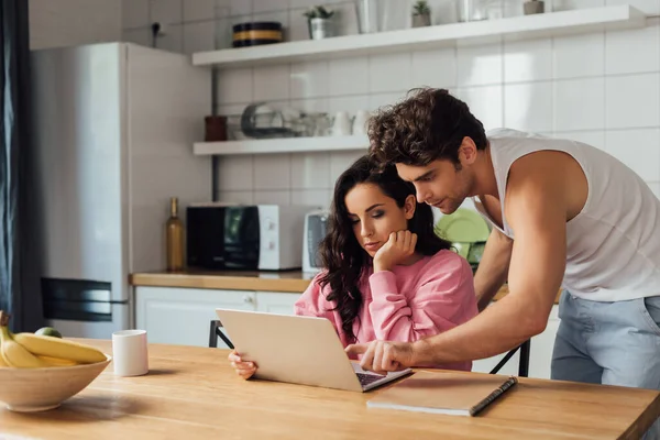 Enfoque selectivo del hombre apuntando con el dedo a la computadora portátil cerca de la novia y portátil en la encimera de la cocina — Stock Photo