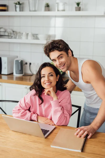 Enfoque selectivo de pareja joven positiva mirando la cámara cerca de portátil y portátil en la mesa de la cocina - foto de stock