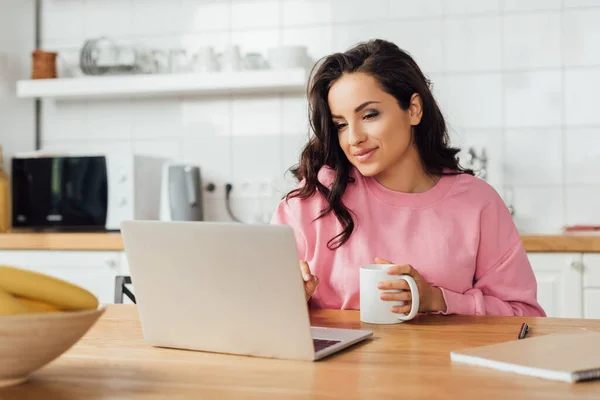 Focus selettivo di bella donna in possesso di una tazza di caffè vicino al computer portatile e notebook sul tavolo della cucina — Foto stock