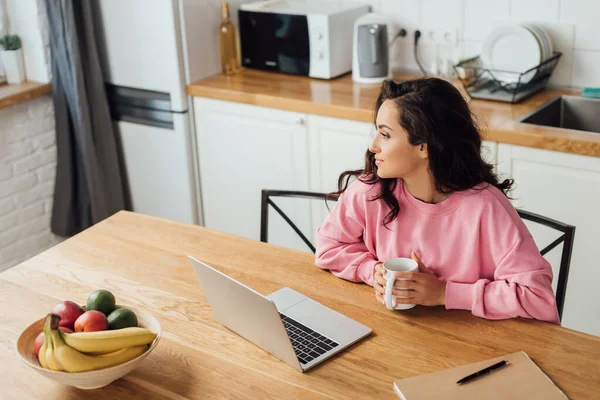 Vista ad alto angolo di giovane donna che tiene una tazza di caffè vicino a laptop, notebook e frutta fresca sul tavolo della cucina — Foto stock