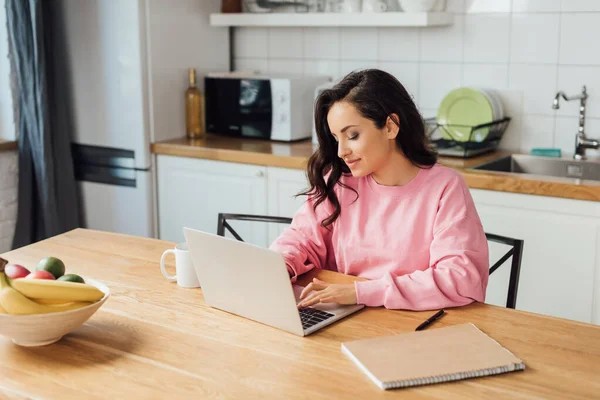 Selective focus of beautiful freelancer using laptop near notebook and cup of coffee in kitchen — Stock Photo