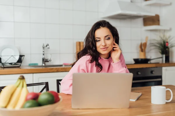 Selective focus of attractive freelancer looking at laptop near notebook, cup and fresh fruits in kitchen — Stock Photo