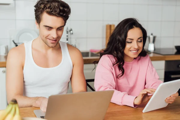 Selektiver Fokus eines lächelnden Paares mit Laptop und digitalem Tablet auf dem Küchentisch — Stockfoto