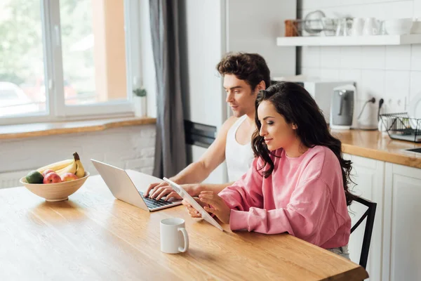 Селективный фокус молодой женщины с помощью цифрового планшета рядом с парнем с ноутбуком, фруктами и чашкой кофе на кухонном столе — стоковое фото