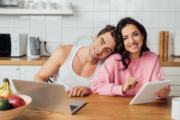 Selektiver Fokus eines Paares, das in die Kamera lächelt, während es Gadgets auf dem Küchentisch benutzt — Stockfoto