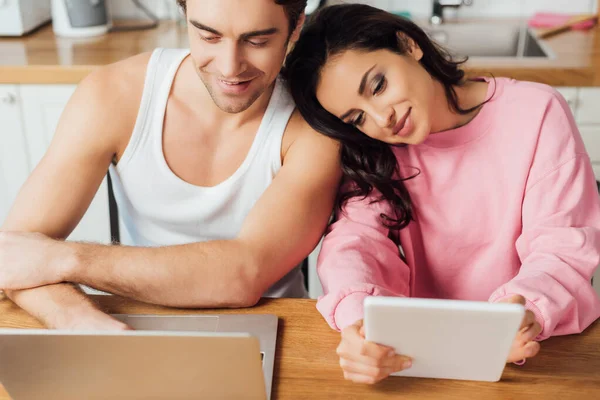 Enfoque selectivo del hombre sonriente utilizando el ordenador portátil cerca de hermosa novia con tableta digital en la cocina - foto de stock