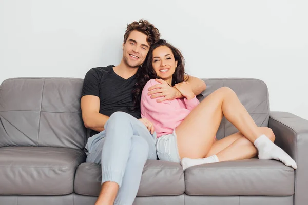 Bonito homem abraçando alegre namorada no sofá no fundo cinza — Fotografia de Stock