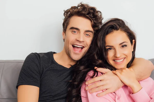 Joven abrazando a su novia sonriente mientras mira a la cámara en el sofá sobre fondo gris - foto de stock