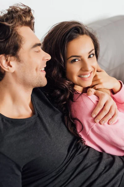 Enfoque selectivo del hombre guapo abrazando a la hermosa mujer sonriente en el sofá sobre fondo gris - foto de stock