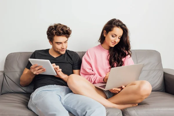 Молодий чоловік тримає цифровий планшет біля усміхненої дівчини, використовуючи ноутбук на дивані на сірому фоні — стокове фото