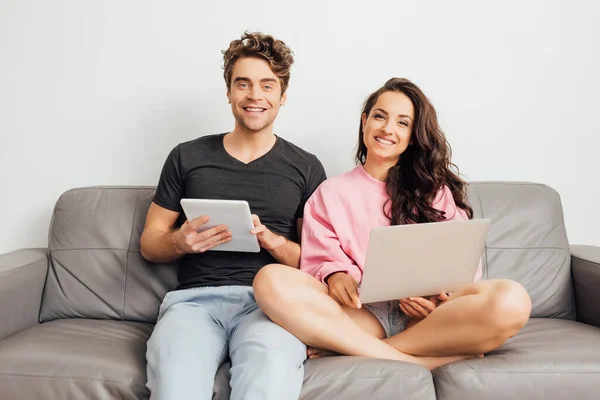 Молодая пара улыбается в камеру, держа ноутбук и цифровой планшет на диване на сером фоне — стоковое фото