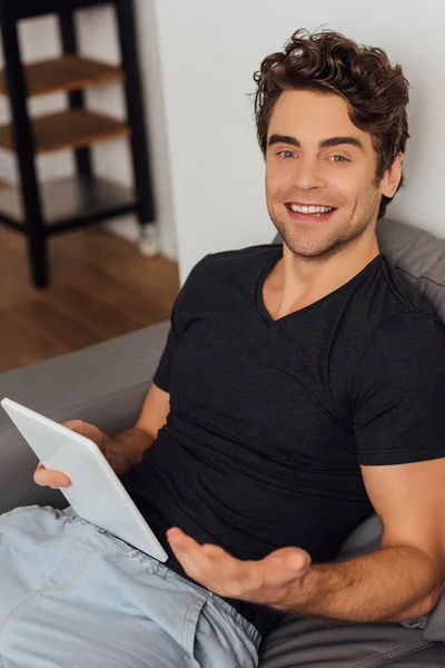 Focus selettivo di bell'uomo che sorride alla fotocamera mentre tiene il tablet digitale sul divano a casa — Foto stock