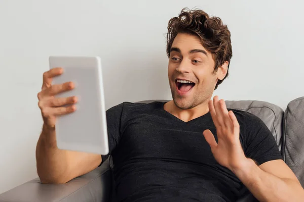 Selektiver Fokus eines fröhlichen Mannes, der mit der Hand wedelt, während er auf der Couch auf grauem Hintergrund ein digitales Tablet anruft — Stockfoto