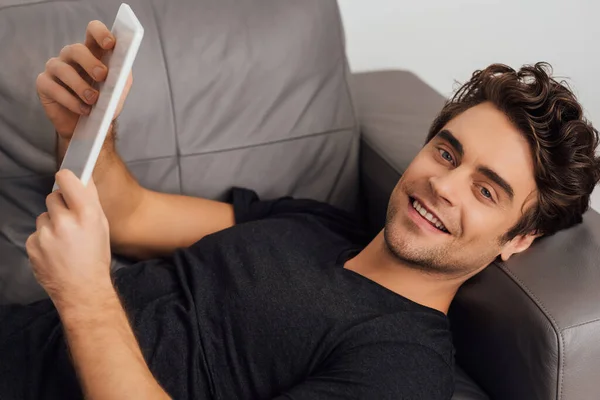 Homme souriant regardant la caméra tout en tenant tablette numérique sur le canapé isolé sur gris — Photo de stock