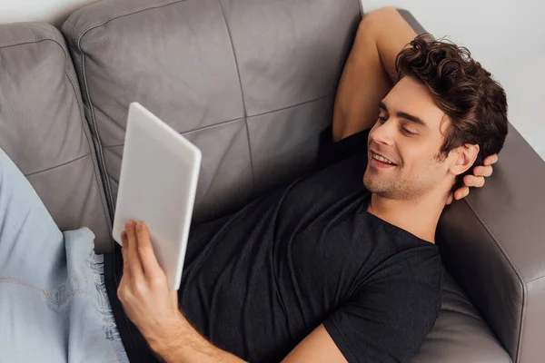 Selektiver Fokus eines gutaussehenden Mannes, der lächelnd ein digitales Tablet auf dem Sofa vor grauem Hintergrund hält — Stockfoto