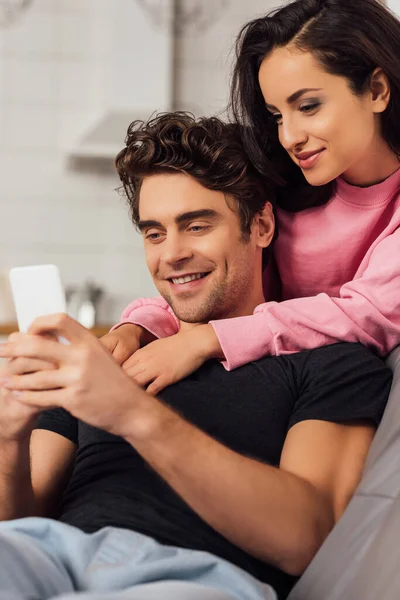 Enfoque selectivo de la hermosa mujer abrazando novio sonriente utilizando el teléfono inteligente en casa - foto de stock