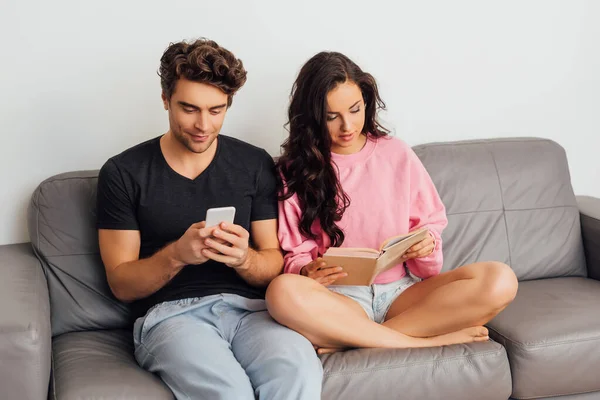 Jovem leitura livro perto namorado usando smartphone no sofá no fundo cinza — Fotografia de Stock