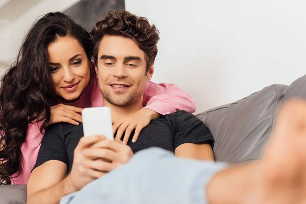 Focus selettivo di uomo sorridente utilizzando smartphone vicino bella fidanzata a casa — Foto stock