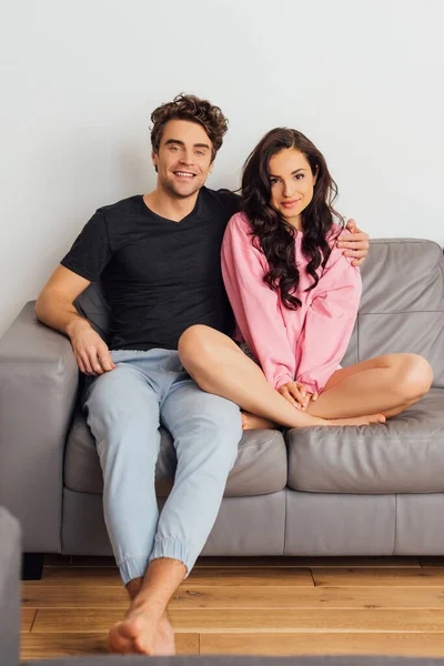 Selektiver Fokus eines lächelnden Mannes, der seine schöne Freundin umarmt und zu Hause auf dem Sofa in die Kamera schaut — Stockfoto
