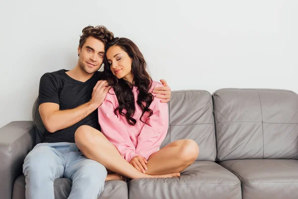 Hombre guapo abrazando a mujer hermosa con los ojos cerrados en sofá sobre fondo gris - foto de stock