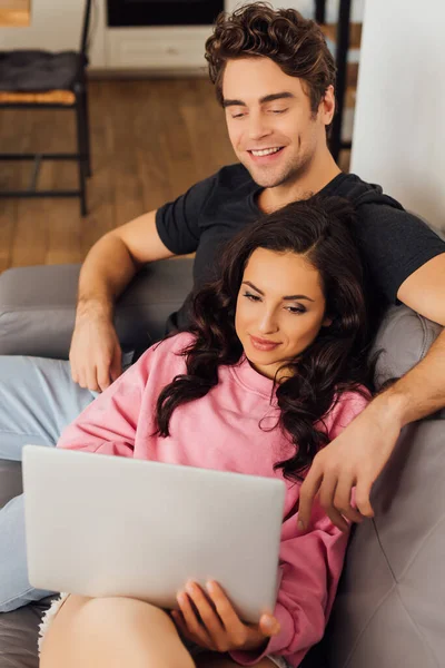 Selective focus of smiling man sitting near girlfriend using laptop on sofa at home — Stock Photo