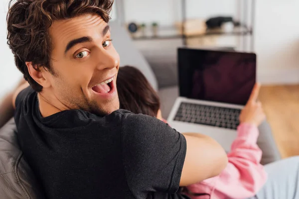 Selektiver Fokus eines fröhlichen Mannes, der in die Kamera schaut, während seine Freundin zu Hause den Laptop benutzt — Stockfoto