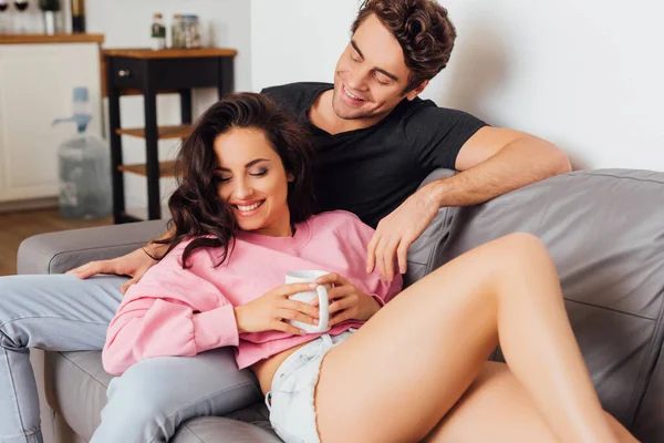 Lächelnde Frau hält Tasse Kaffee auf Couch neben Freund im Wohnzimmer — Stockfoto