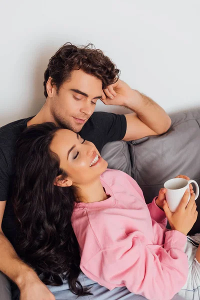 Красивый мужчина сидит рядом с веселой улыбающейся девушкой, держа чашку на диване — стоковое фото