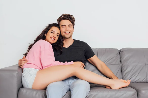 Joven abrazando a su novia y sonriendo a la cámara en el sofá aislado en gris - foto de stock