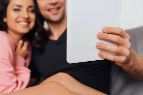 Enfoque selectivo del hombre sosteniendo la tableta digital cerca de la novia alegre en el sofá aislado en gris - foto de stock