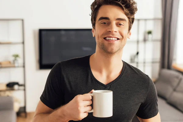 Hombre alegre sosteniendo una taza de café y sonriendo a la cámara en casa - foto de stock