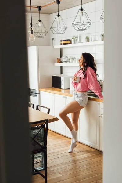 Selective focus of beautiful smiling girl holding cup of coffee in kitchen — Stock Photo