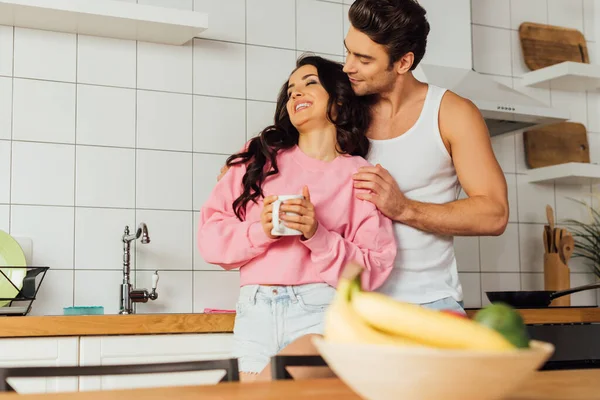 Foco seletivo do homem abraçando bela namorada sorridente com xícara de café na cozinha — Fotografia de Stock