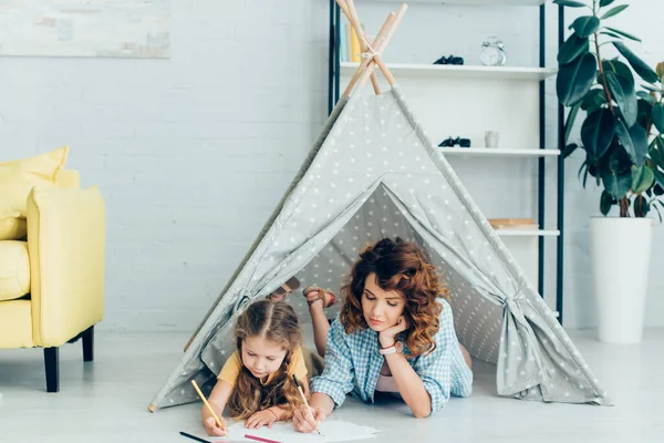 Young nanny and cute kid drawing together while lying in toy wigwam — Stock Photo