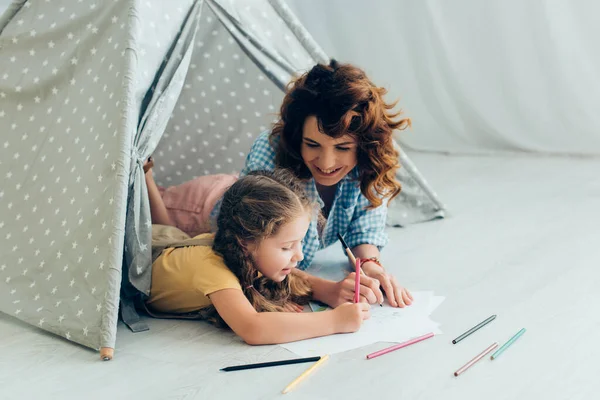 Улыбающаяся няня и очаровательный ребенок рисует вместе, лежа в игровой палатке — стоковое фото