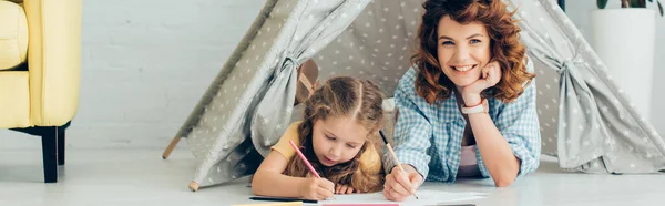 Happy nanny looking at camera while drawing together with child in play tent, panoramic shot — Stock Photo