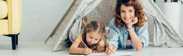 Glückliche Kindermädchen blickt in die Kamera, während sie mit Kind im Spielzelt zeichnet, Panoramaaufnahme — Stockfoto