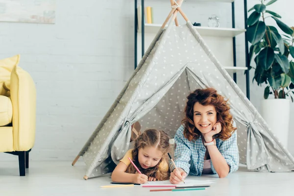 Happy nanny looking at camera while drawing together with cute child in kids wigwam — Stock Photo