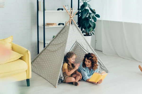Sorridente babá leitura livro para adorável criança em jogar tenda — Fotografia de Stock
