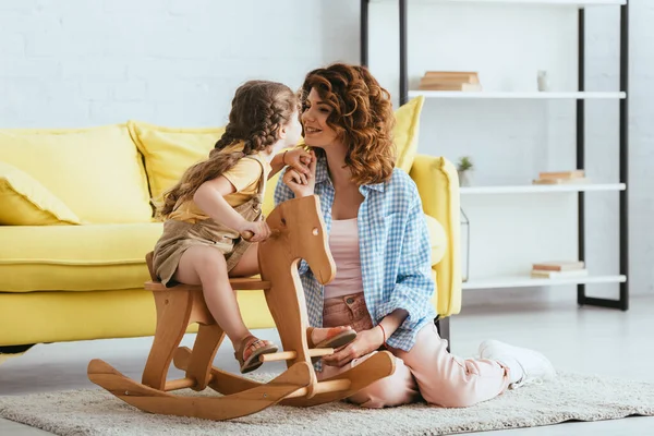Happy nanny sitting on floor and holding hand of child riding rocking horse — Stock Photo